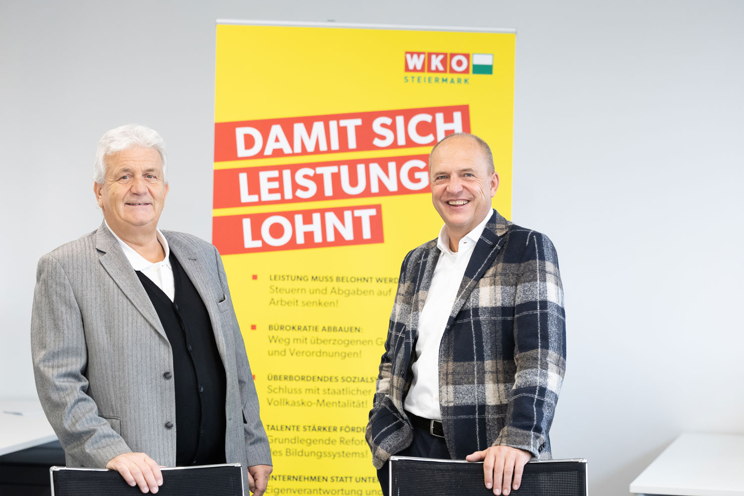 Zwei lächelnde Männer in Geschäftskleidung stehen vor einem großen gelb-roten Plakat der WKO Steiermark mit der Aufschrift "Damit sich Leistung lohnt" in einer hellen Büroumgebung.