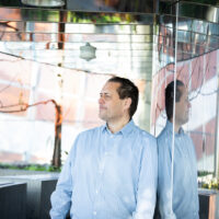 Ein Mann in einem hellblauen Hemd steht in einem Innenraum und lehnt sich gegen eine verspiegelte Wand, in der sein Spiegelbild zu sehen ist. Im Hintergrund sind moderne Architektur, Glas, Pflanzen und natürliches Licht zu sehen.