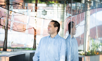 Ein Mann in einem hellblauen Hemd steht in einem Innenraum und lehnt sich gegen eine verspiegelte Wand, in der sein Spiegelbild zu sehen ist. Im Hintergrund sind moderne Architektur, Glas, Pflanzen und natürliches Licht zu sehen.