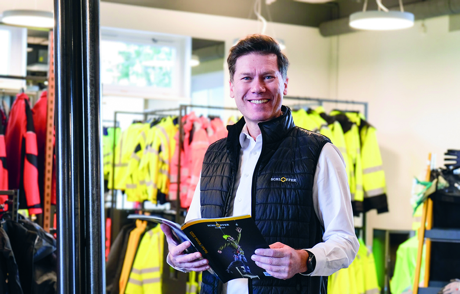 Ein lächelnder Mann in schwarzer Weste und weißem Hemd steht in einem Raum und hält eine Zeitschrift in der Hand. Hinter ihm stehen in einem hell erleuchteten Raum Regale mit Warnschutzjacken und Arbeitskleidung.