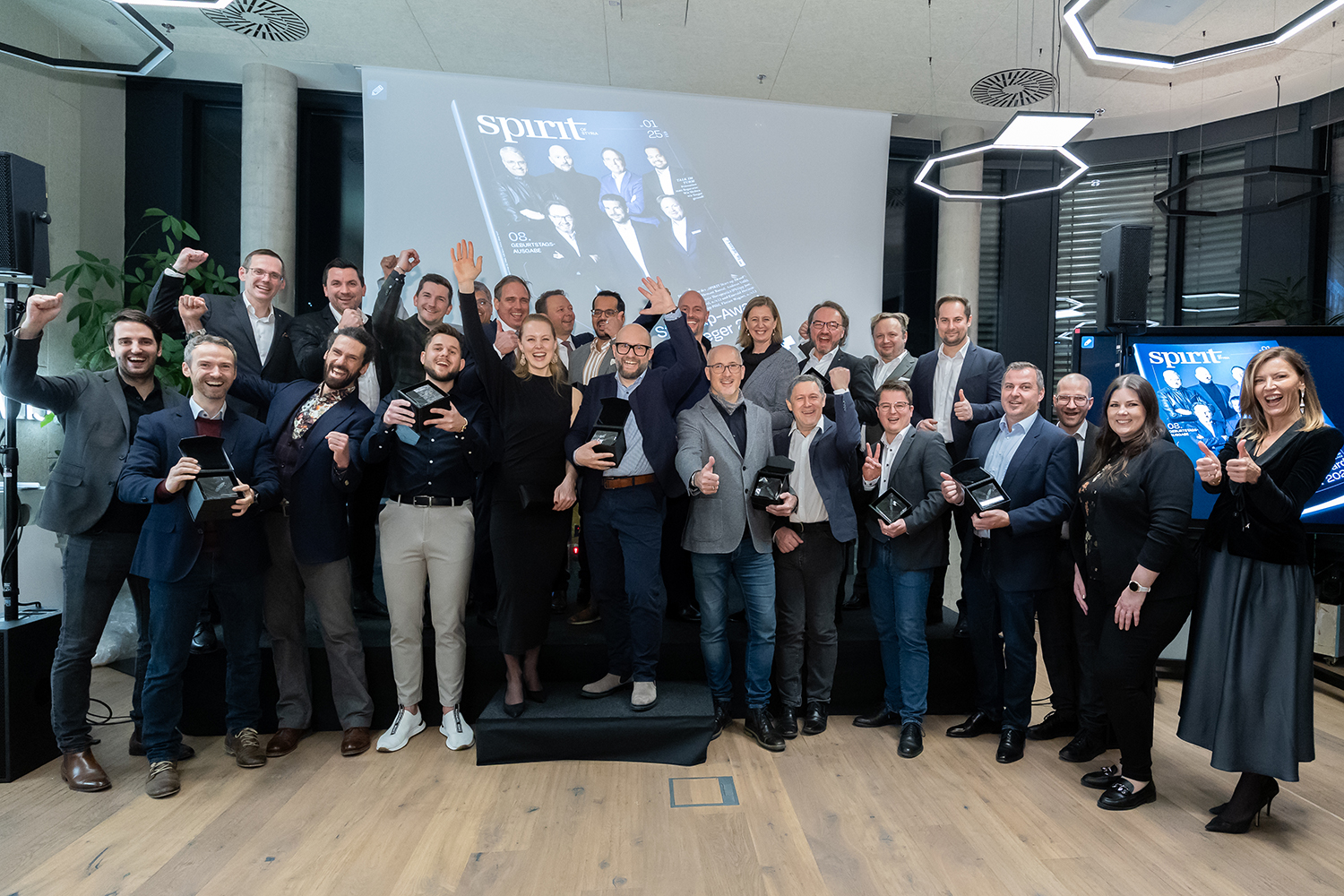 Eine große Gruppe von Menschen in Geschäftskleidung steht in einem Innenraum zusammen, lächelt und posiert für ein Festtagsfoto. Einige halten Trophäen, und im Hintergrund ist ein projiziertes Zeitschriftencover zu sehen.