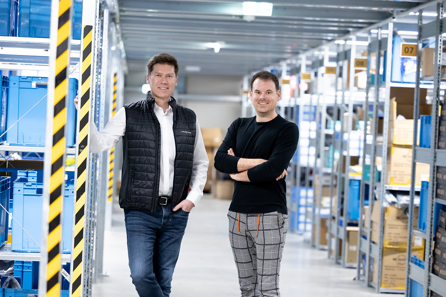 Zwei Männer stehen lächelnd in einem gut beleuchteten Lagergang voller Regale und blauer Lagerkästen. Ein Mann trägt eine schwarze Weste und Jeans, der andere einen schwarzen Pullover und eine karierte Hose.