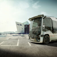 Ein moderner, futuristischer, gläserner Bus mit der Aufschrift "Harlander" steht an einem hellen, klaren Tag auf einer leeren Straße in der Nähe moderner Glasgebäude.