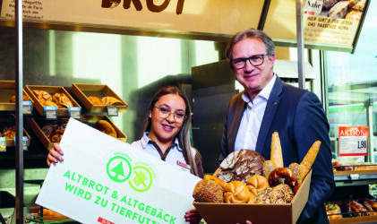 Eine Frau und ein Mann stehen lächelnd in einer Bäckerei. Die Frau hält ein Schild mit der Aufschrift "Altbrot & Altgebäck wird zu Tierfutter", und der Mann hält einen Korb mit verschiedenen Brotsorten und Brötchen. Im Hintergrund sind Brotregale zu sehen.