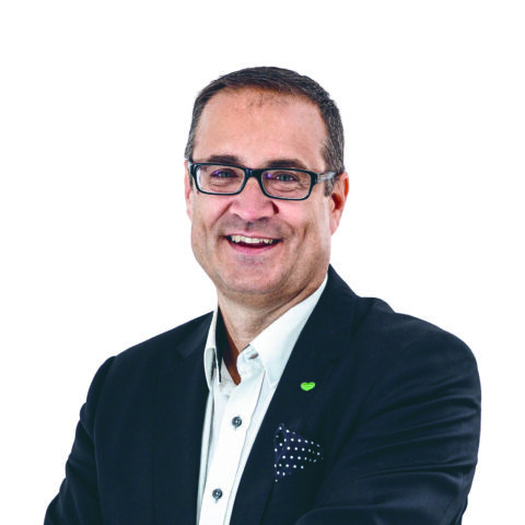 Ein Mann mit kurzen dunklen Haaren und Brille lächelt, während er eine schwarze Anzugsjacke, ein weißes Hemd und ein gepunktetes Einstecktuch trägt und vor einem schlichten weißen Hintergrund steht.