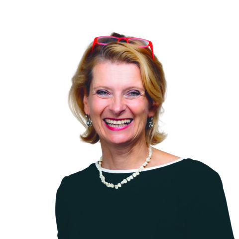 Lächelnde Frau mit kurzen blonden Haaren, die ein schwarzes Oberteil, eine weiße Perlenkette, Perlenohrringe und eine rote Brille trägt, die auf ihrem Kopf ruht, vor einem weißen Hintergrund.