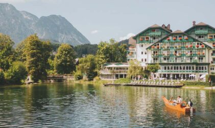 Ein großes, grün gestrichenes Hotel am Seeufer steht in der Nähe des Ufers, umgeben von Bergen und Bäumen. Menschen rudern mit einem Holzboot auf dem ruhigen See unter einem klaren Himmel.