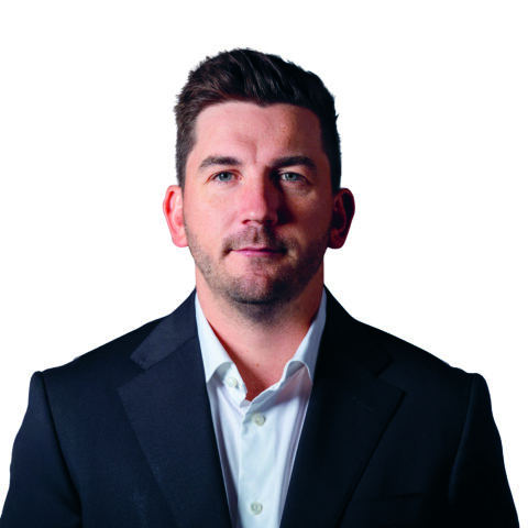 Ein Mann mit kurzem braunem Haar und gestutztem Bart, der ein weißes Hemd und einen dunklen Blazer trägt und vor einem schlichten weißen Hintergrund direkt in die Kamera schaut.