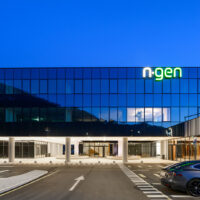 Ein modernes Glasgebäude mit dem beleuchteten Schild "n-gen" im obersten Stockwerk, fotografiert in der Abenddämmerung. Ein silbernes Auto ist davor geparkt, und das Gebäude verfügt über große Fenster und helle Innenbeleuchtung.