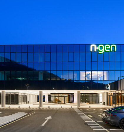 Ein modernes Glasgebäude mit dem beleuchteten Schild "n-gen" im obersten Stockwerk, fotografiert in der Abenddämmerung. Ein silbernes Auto ist davor geparkt, und das Gebäude verfügt über große Fenster und helle Innenbeleuchtung.