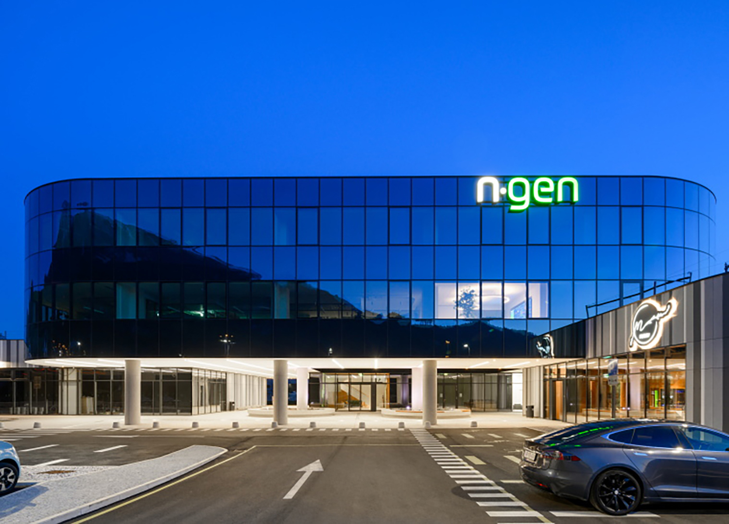 Ein modernes Glasgebäude mit dem beleuchteten Schild "n-gen" im obersten Stockwerk, fotografiert in der Abenddämmerung. Ein silbernes Auto ist davor geparkt, und das Gebäude verfügt über große Fenster und helle Innenbeleuchtung.