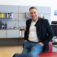 Ein Mann in Blazer und Jeans sitzt an einem roten Schreibtisch in einem modernen Büro und lächelt in die Kamera. Im Hintergrund sind Bücherregale, ein Schreibtisch mit einem Computer, ein Schutzhelm und die Büroeinrichtung zu sehen.