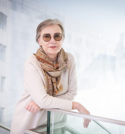 Eine ältere Frau mit kurzen grauen Haaren und einer runden Sonnenbrille lehnt an einem Glasgeländer in einem Innenraum. Sie trägt ein cremefarbenes Outfit und einen gemusterten Schal. Der Hintergrund ist hell und modern mit unscharfen architektonischen Details.
