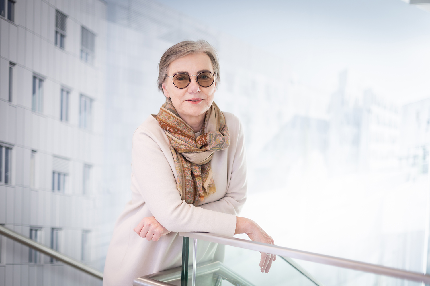 Eine ältere Frau mit kurzen grauen Haaren und einer runden Sonnenbrille lehnt an einem Glasgeländer in einem Innenraum. Sie trägt ein cremefarbenes Outfit und einen gemusterten Schal. Der Hintergrund ist hell und modern mit unscharfen architektonischen Details.