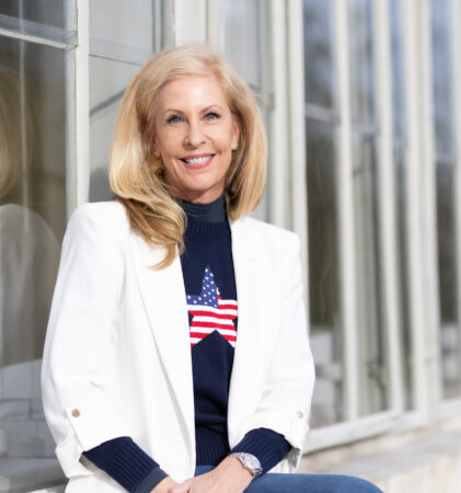 Eine lächelnde Frau mit langen blonden Haaren, die einen weißen Blazer und einen marineblauen Pullover mit der amerikanischen Flagge trägt, sitzt an einem sonnigen Tag vor einem großen Fenster.
