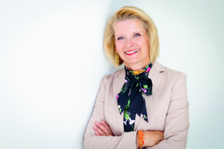 Eine lächelnde ältere Frau mit kurzen blonden Haaren steht vor einem weißen Hintergrund. Sie trägt einen hellbeigen Blazer, ein buntes Halstuch und goldene Ohrringe. Ihre Arme sind verschränkt, und sie sieht selbstbewusst und freundlich aus.