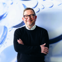 Ein Mann mit Brille und Bart steht lächelnd mit verschränkten Armen vor einem blau-weißen abstrakten Hintergrund mit Kreisformen und Blasen.