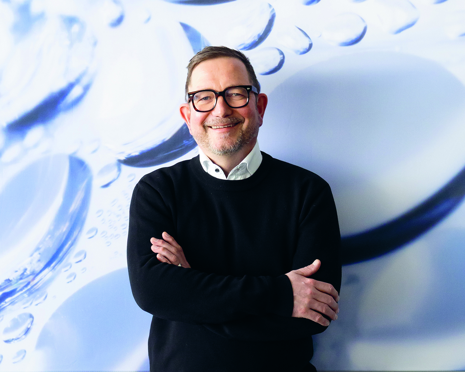 Ein Mann mit Brille und Bart steht lächelnd mit verschränkten Armen vor einem blau-weißen abstrakten Hintergrund mit Kreisformen und Blasen.