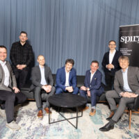 Sieben Männer in Geschäftskleidung posieren und lächeln in einem modernen Raum mit einem blauen Vorhang im Hintergrund, sitzend und stehend um einen schwarzen Couchtisch, mit einem "Spirit"-Banner im Hintergrund.