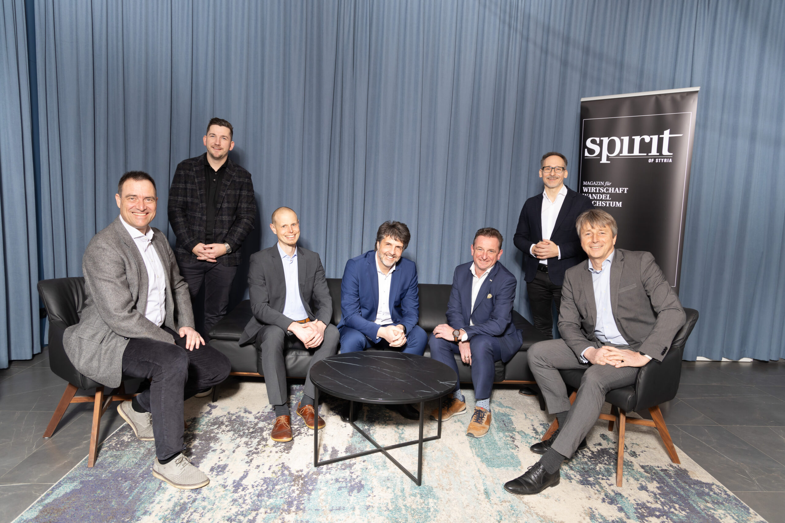 Sieben Männer in Geschäftskleidung posieren und lächeln in einem modernen Raum mit einem blauen Vorhang im Hintergrund, sitzend und stehend um einen schwarzen Couchtisch, mit einem "Spirit"-Banner im Hintergrund.