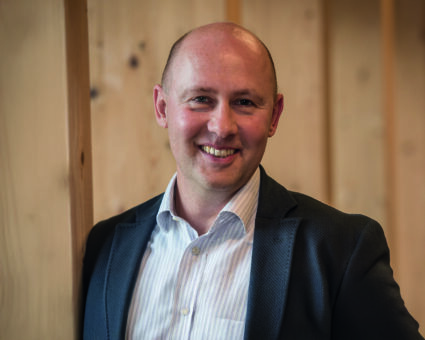 Ein lächelnder Mann in Blazer und gestreiftem Hemd lehnt an einer Holzwand und schaut direkt in die Kamera. Im Hintergrund sind vertikale Holzpaneele zu sehen.