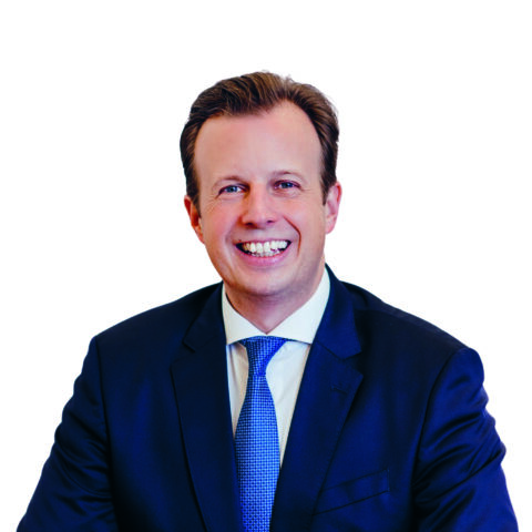 Ein Mann in einem dunklen Anzug und mit blauer Krawatte lächelt in einem Kopf-Schulter-Porträt vor weißem Hintergrund.