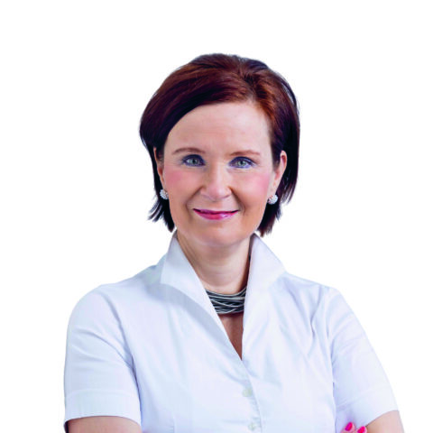 Eine Frau mit kurzen braunen Haaren, die ein weißes Hemd mit Kragen und Silberschmuck trägt, lächelt selbstbewusst mit verschränkten Armen vor einem schlichten weißen Hintergrund.