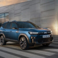 Ein blauer Dacia Geländewagen fährt auf einer Stadtstraße in der Nähe eines modernen Gebäudes mit spiegelnden Glasfenstern und einer strukturierten Betonwand unter einem leicht bewölkten Himmel.