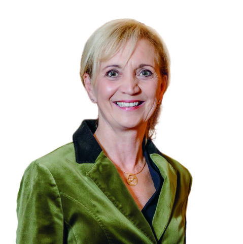 Eine lächelnde Frau mittleren Alters mit kurzem blondem Haar, die einen grünen Samtblazer mit dunklem Kragen und eine goldene Halskette trägt, posiert vor einem weißen Hintergrund.