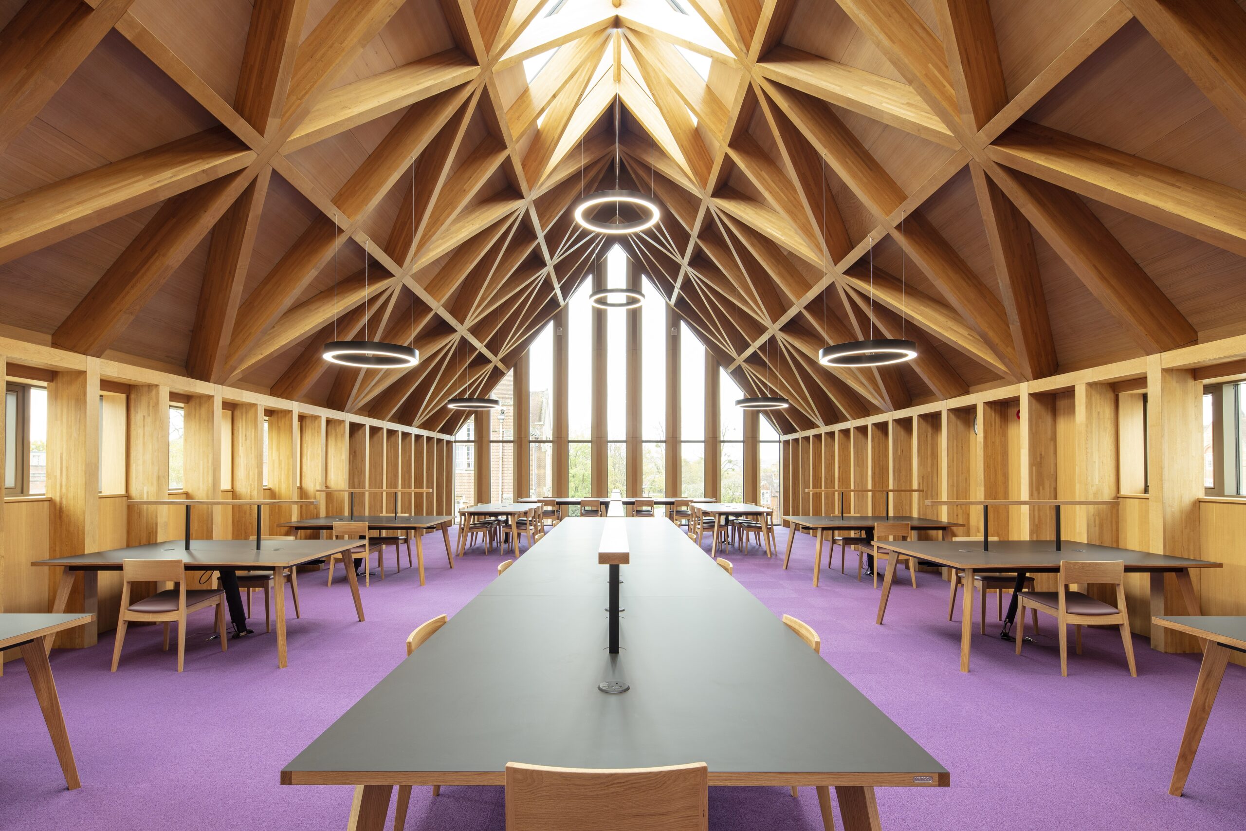 Eine geräumige Bibliothek oder ein Arbeitszimmer mit einer hohen, hölzernen Gewölbedecke, runden Hängelampen, großen Fenstern und Reihen von Tischen und Stühlen auf einem hellvioletten Teppich.