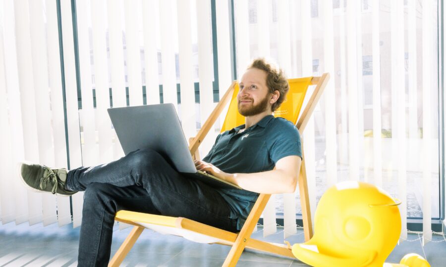 Ein Mann sitzt entspannt in einem Liegestuhl in einem Haus und benutzt einen Laptop auf seinem Schoß. Er lächelt und steht in der Nähe einer großen, gelben Gummiente. Weiße vertikale Jalousien verdecken die Fenster hinter ihm und lassen natürliches Licht herein.