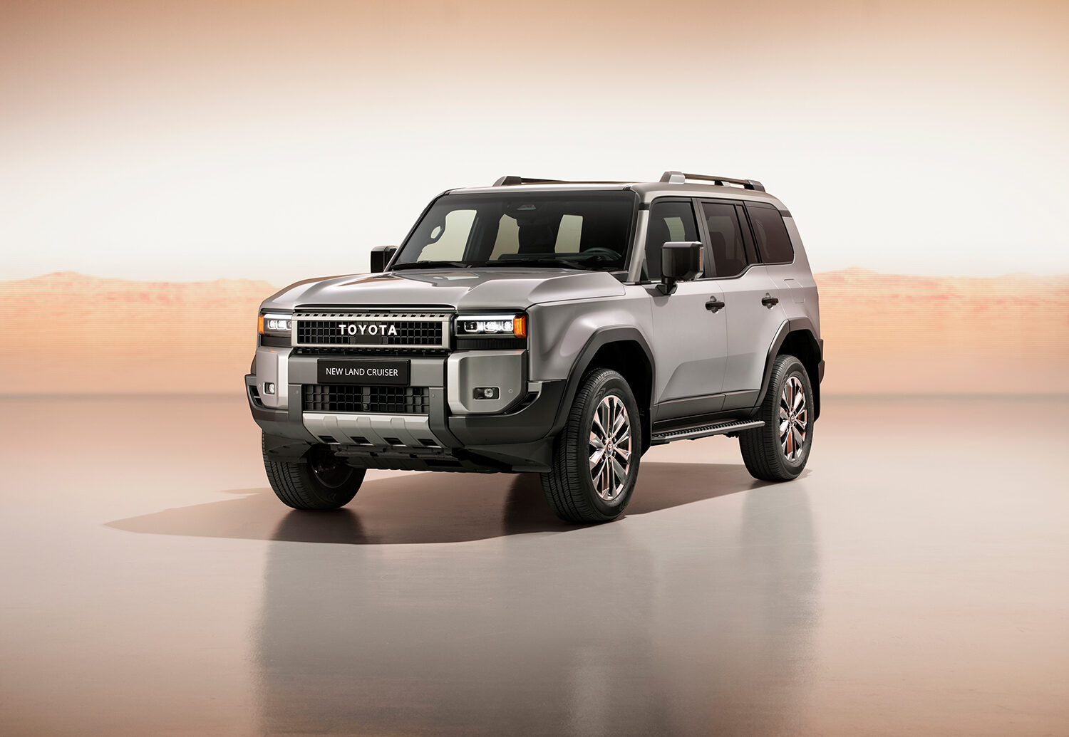Ein silberner Toyota Land Cruiser Geländewagen wird in einem hell beleuchteten Innenraum mit einer Wüstenlandschaft im Hintergrund gezeigt. Das Fahrzeug ist leicht nach links ausgerichtet, was sein modernes und robustes Design unterstreicht.