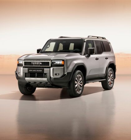 Ein silberner Toyota Land Cruiser Geländewagen wird in einem hell beleuchteten Innenraum mit einer Wüstenlandschaft im Hintergrund gezeigt. Das Fahrzeug ist leicht nach links ausgerichtet, was sein modernes und robustes Design unterstreicht.