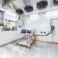 Helles Labor mit einem Metallarbeitsplatz, Computern, Kabeln und wissenschaftlichen Geräten. An der Wand montierte Ventilatoren und elektrische Schalttafeln sind sichtbar. Der Raum hat saubere, weiße Wände und ein Fenster, das in einen anderen Raum führt.