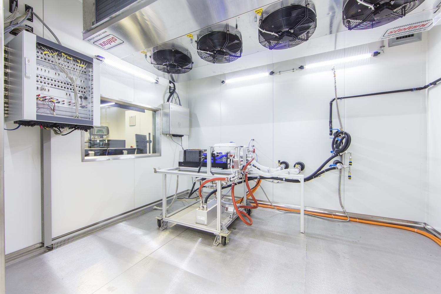 Helles Labor mit einem Metallarbeitsplatz, Computern, Kabeln und wissenschaftlichen Geräten. An der Wand montierte Ventilatoren und elektrische Schalttafeln sind sichtbar. Der Raum hat saubere, weiße Wände und ein Fenster, das in einen anderen Raum führt.