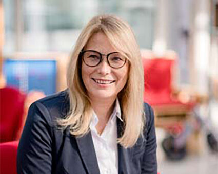 Eine Frau mit glattem blondem Haar und Brille, die einen dunklen Blazer über einem weißen Hemd trägt, lächelt, während sie in einem Raum mit roten Stühlen und einem hellen, verschwommenen Hintergrund sitzt.