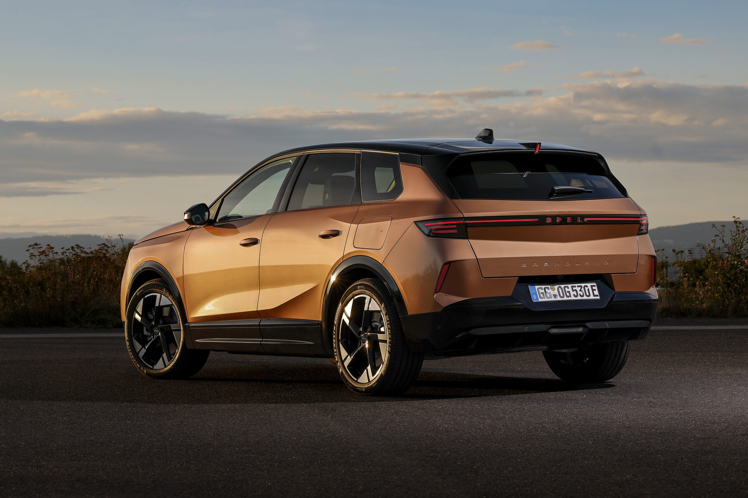 Ein bronzener Opel Grandland SUV ist bei Sonnenuntergang auf einer Straße geparkt, mit bewölktem Himmel und einer malerischen Landschaft im Hintergrund. Das Auto ist aus der hinteren Dreiviertelperspektive zu sehen.