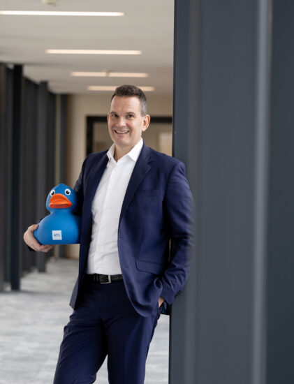 Ein Mann in einem dunkelblauen Anzug steht in einem modernen Flur, lächelt und hält eine große blaue Gummiente unter seinem Arm.