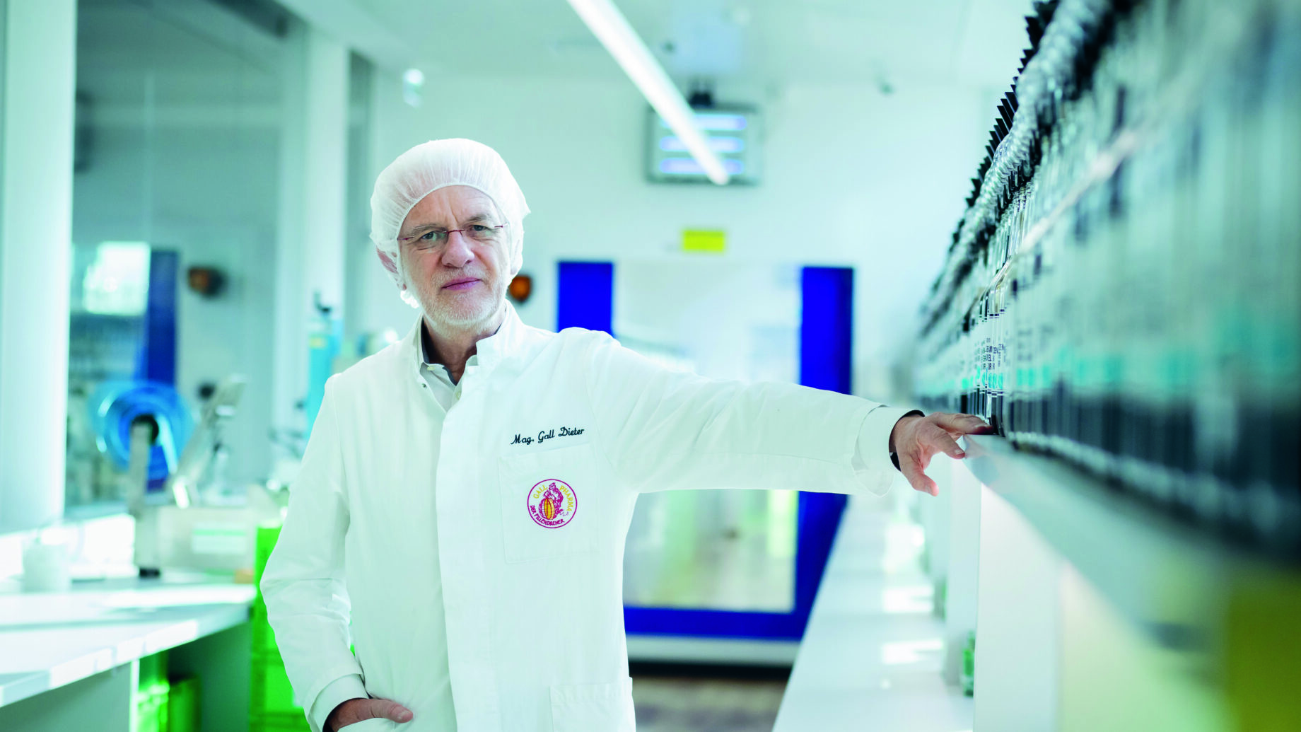 Ein Wissenschaftler in weißem Laborkittel und Haarnetz steht in einem hellen Labor, eine Hand stützt sich auf ein Regal mit Flaschen. Im Hintergrund sind Laborgeräte und eine blaue Tür zu sehen.
