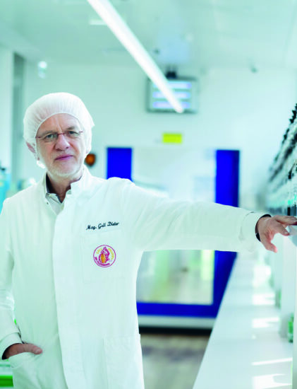 Ein Wissenschaftler in weißem Laborkittel und Haarnetz steht in einem hellen Labor, eine Hand stützt sich auf ein Regal mit Flaschen. Im Hintergrund sind Laborgeräte und eine blaue Tür zu sehen.