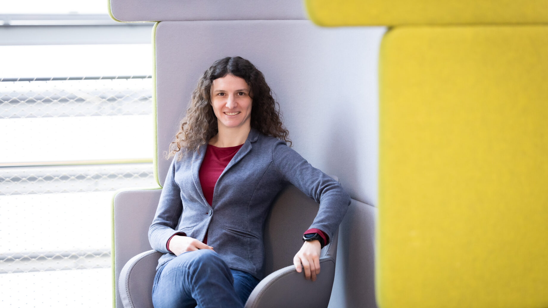Eine Person mit langen lockigen Haaren, die einen blauen Blazer und Jeans trägt, sitzt lächelnd auf einem modernen Stuhl neben hellgrauen und gelben Wandpaneelen in einem hellen Raum.