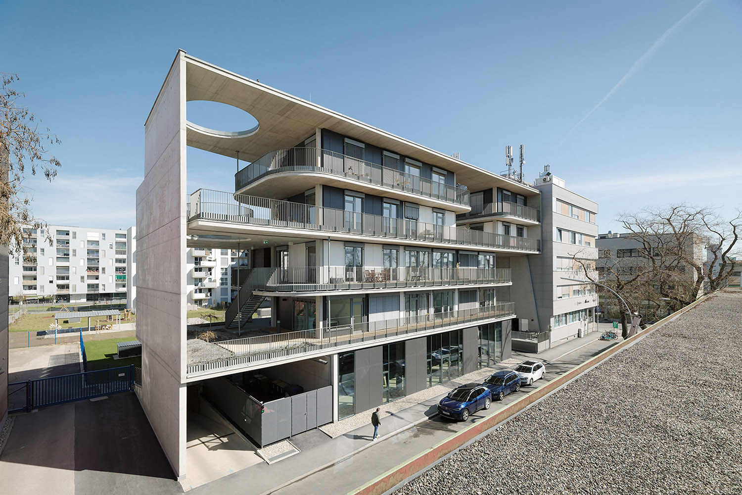 Ein modernes mehrstöckiges Gebäude mit großen Glasbalkonen, geschwungenen architektonischen Elementen und einem runden Ausschnitt im Dach. Vor dem Gebäude sind mehrere Autos geparkt, und eine Person geht unter freiem Himmel auf dem Gehweg entlang.