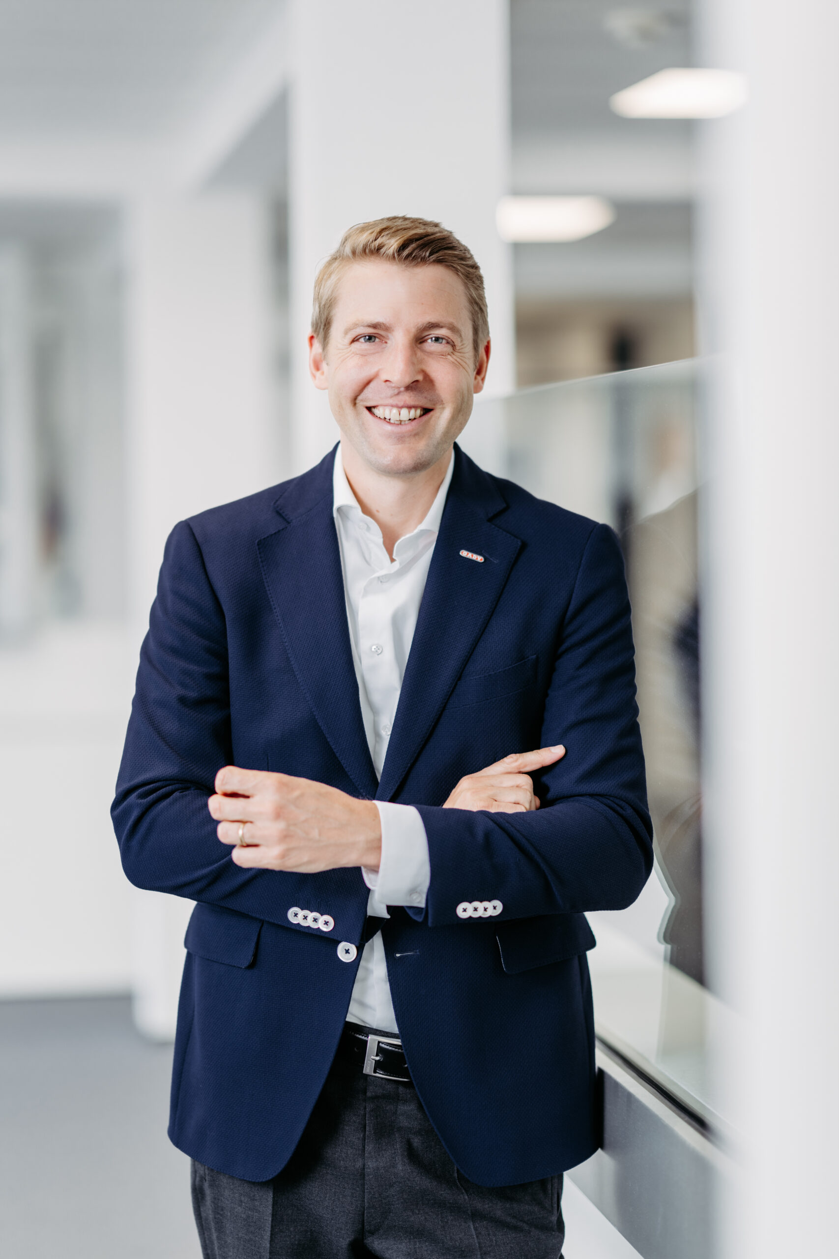 Ein Mann in einem marineblauen Blazer und einem weißen Hemd steht mit verschränkten Armen in einem Haus und lächelt in die Kamera. Der Hintergrund ist hell und modern, mit unscharfen Büroelementen.