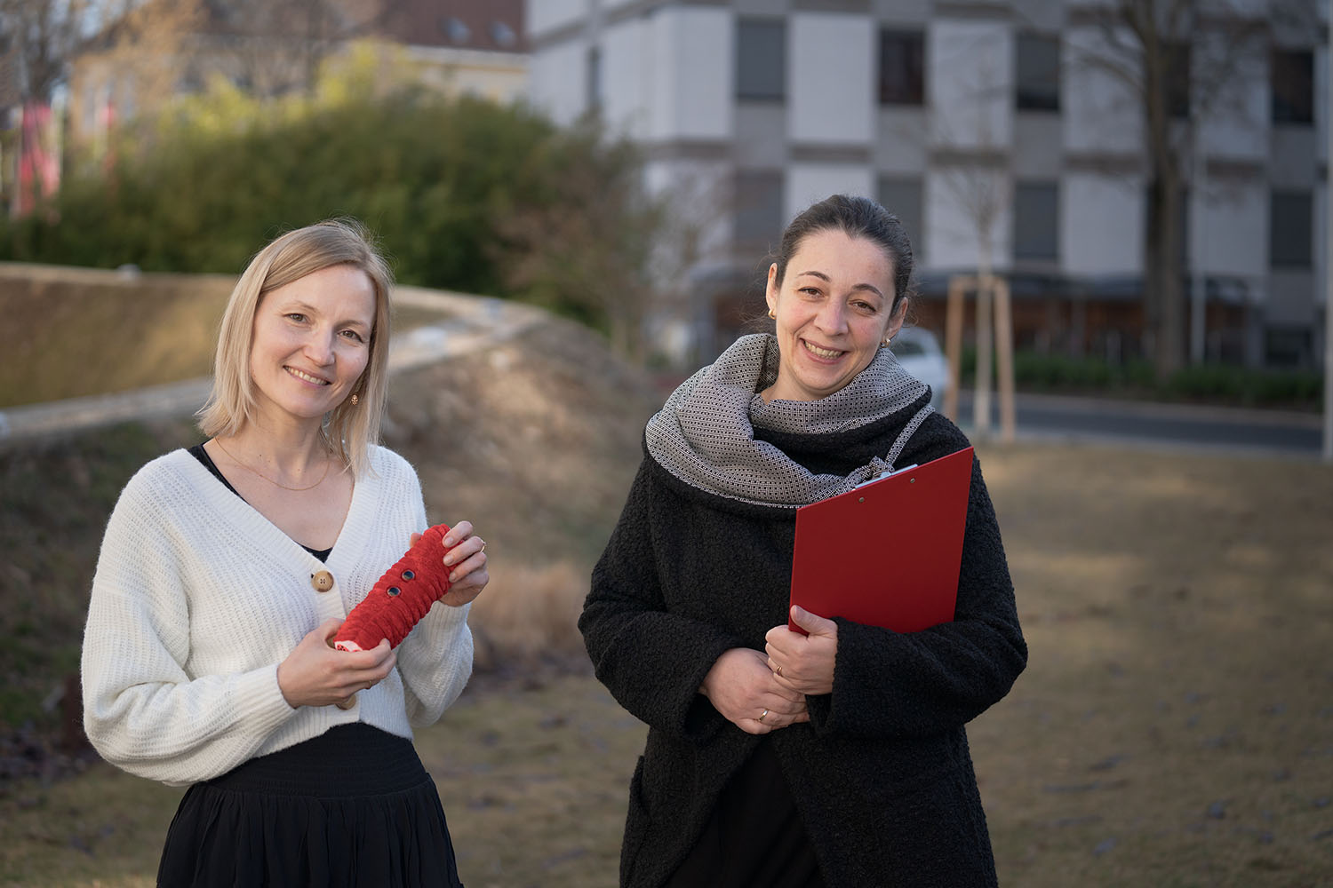 Zwei Frauen stehen im Freien, beide lächeln. Die eine hält ein rotes gehäkeltes Objekt, die andere ein rotes Klemmbrett in der Hand. Im Hintergrund sind Wohnhäuser und kahle Bäume zu sehen.