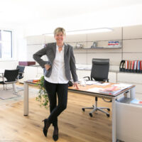 Eine lächelnde Frau in Geschäftskleidung steht in einem modernen, hellen Büro und lehnt an einem Schreibtisch mit geordneten Papieren, Stühlen, Regalen und einem Fenster im Hintergrund.