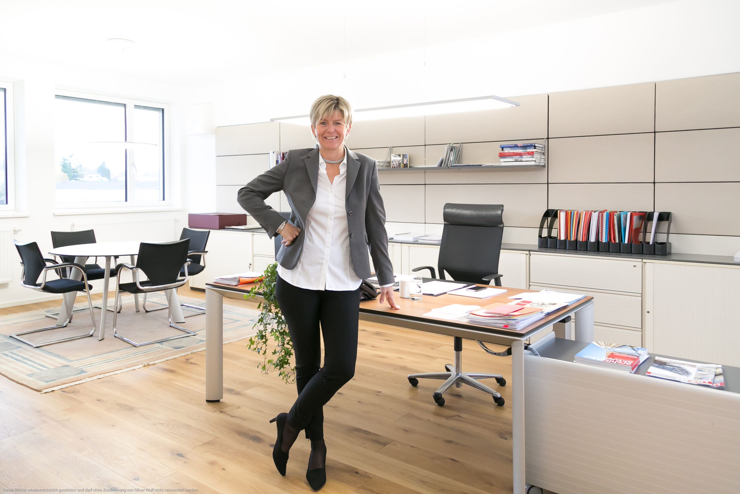 Eine lächelnde Frau in Geschäftskleidung steht in einem modernen, hellen Büro und lehnt an einem Schreibtisch mit geordneten Papieren, Stühlen, Regalen und einem Fenster im Hintergrund.