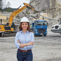 Eine Frau, die einen weißen Schutzhelm und ein blaues Hemd trägt, steht lächelnd mit verschränkten Armen auf einer Baustelle, auf der im Hintergrund Bagger, ein Lastwagen und schwere Maschinen arbeiten.