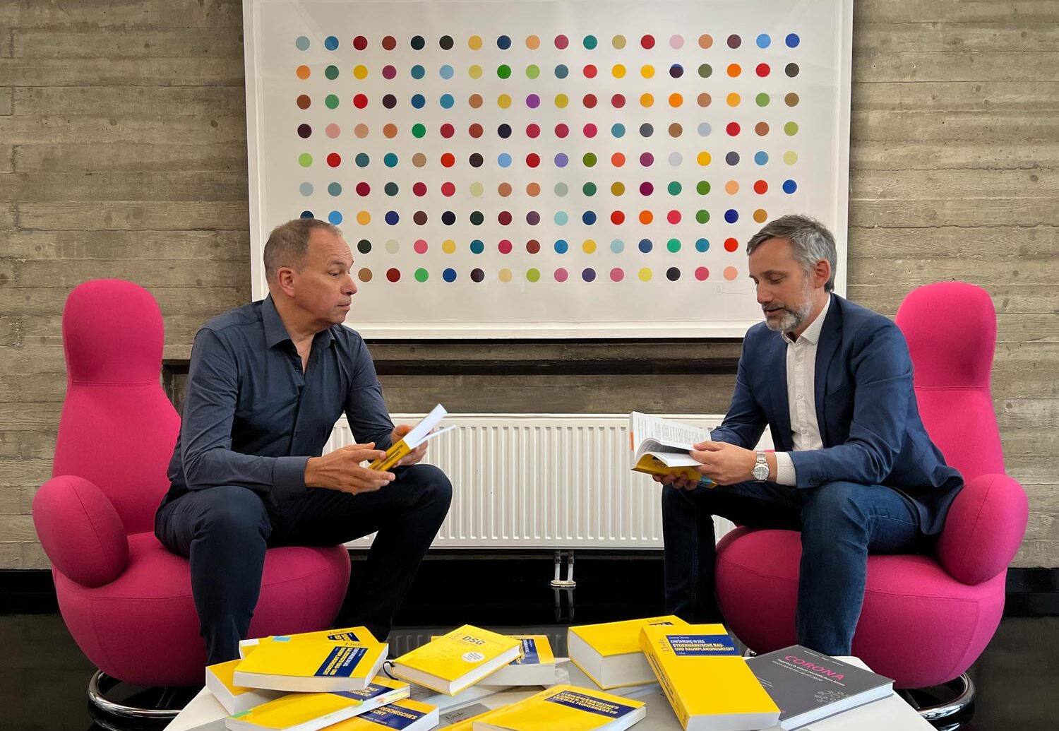 Zwei Männer sitzen sich auf rosa Stühlen gegenüber und lesen Bücher. Vor ihnen steht ein Tisch mit mehreren gelben Büchern. Hinter ihnen ist ein bunt gepunktetes Kunstwerk an einer Betonwand zu sehen.