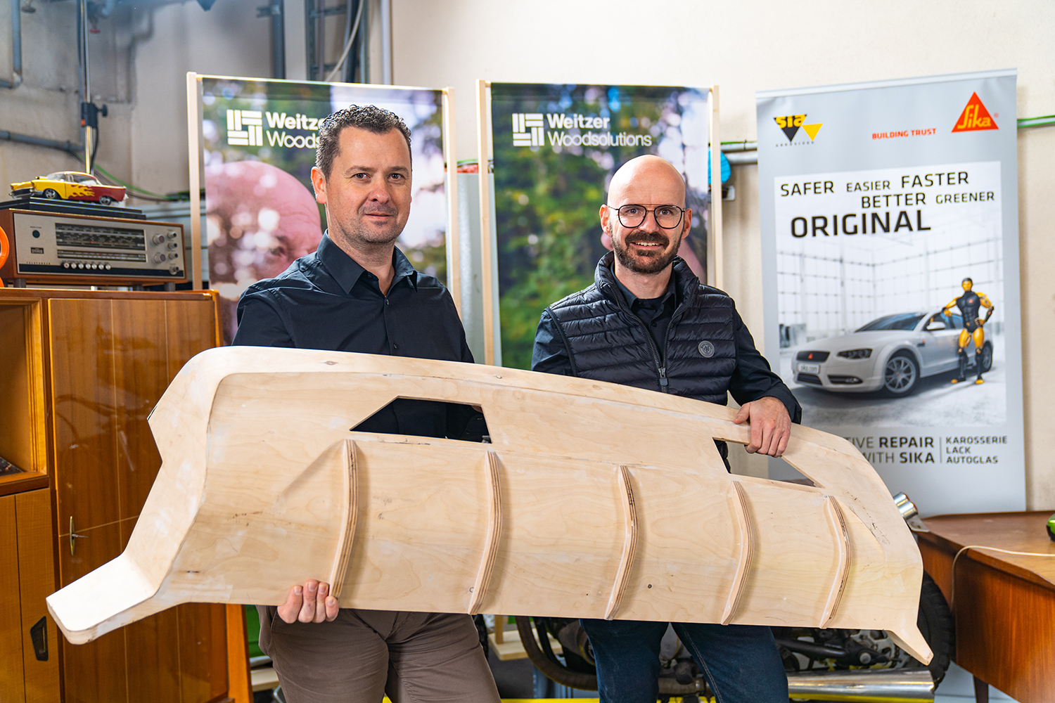 Zwei Männer stehen in einem Haus und halten ein großes Holzmodell eines Autos. Hinter ihnen sind Banner mit Werbung für Autoreparaturen und Firmenlogos zu sehen. Der Raum ist mit Holzschränken und verschiedenen Gegenständen im Hintergrund ausgestattet.