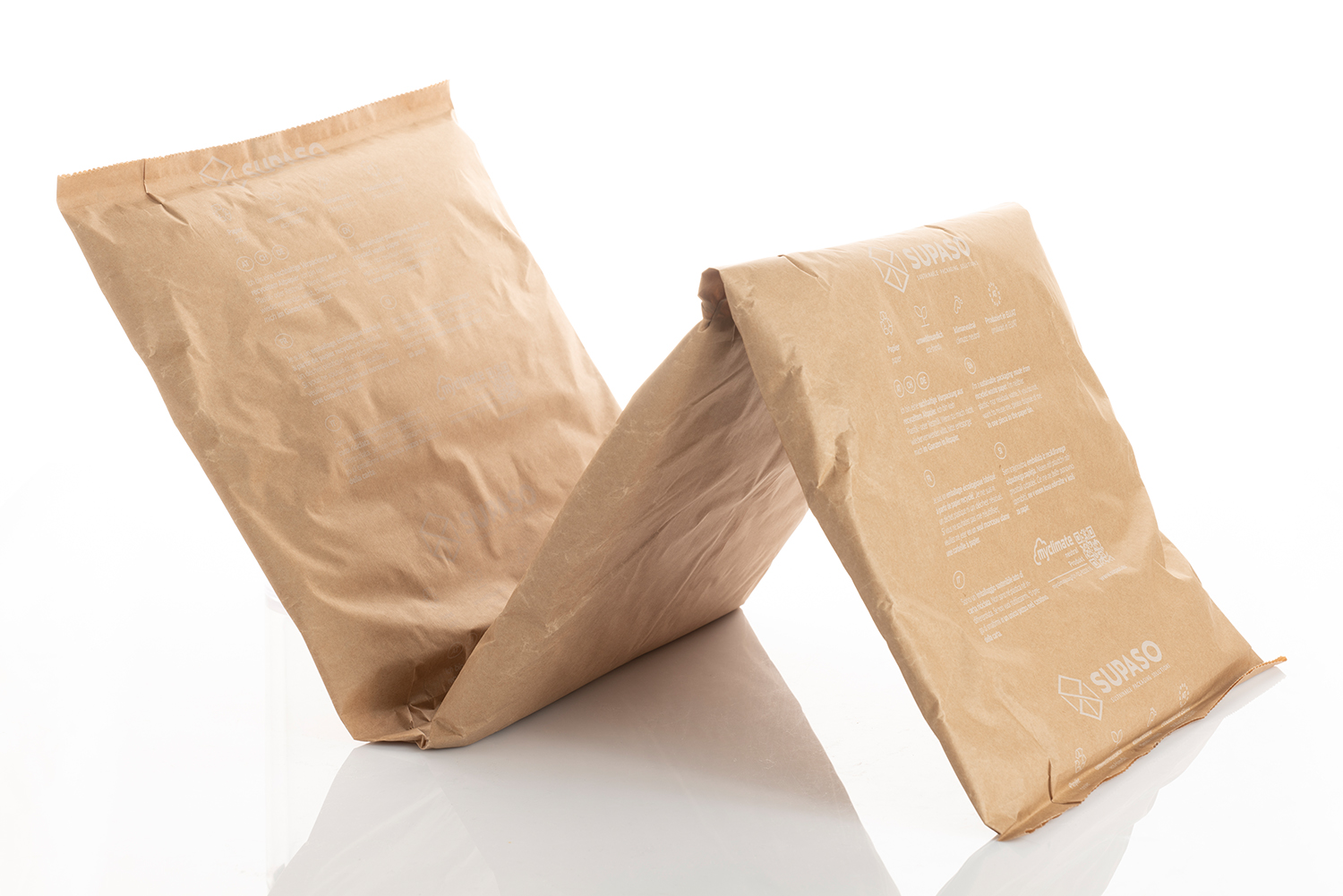 Zwei große gepolsterte Umschläge aus braunem Papier, einer aufrecht, der andere dagegen gelehnt, auf einer weißen, reflektierenden Fläche ausgestellt. Beide Umschläge sind mit weißem Text und Logos bedruckt.